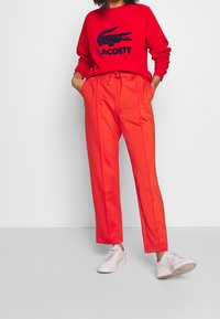 Rode sweatshirt met een marineblauw krokodillenlogo en de tekst "LACOSTE," gecombineerd met felrode tapered pants en witte sneakers.
