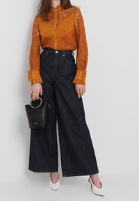 Camicetta in pizzo arancione e raso con dettagli a bottone, abbinata a pantaloni in denim scuro a gamba larga, tacchi bianchi e una borsa nera con manico circolare.