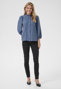 Hellblaue Bluse mit Puffärmeln, Knopfleiste, Rüschenausschnitt, kombiniert mit schwarzen blickdichten Hosen und schwarzen High Heels. Schlicht, strukturiertes Material.