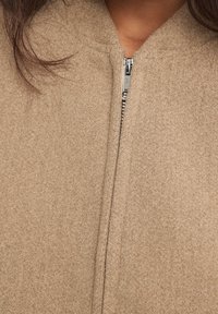 Primer plano de un abrigo de lana beige parcialmente cerrado, usado por una persona con el cabello oscuro visible en los hombros.