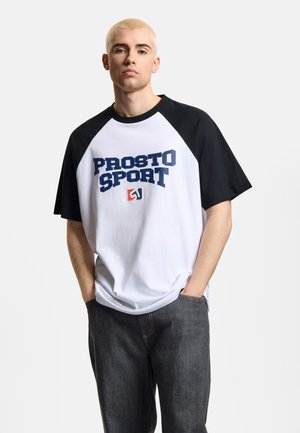 Młody mężczyzna z krótkimi, wybielonymi włosami, ubrany w białą i czarną koszulkę "PROSTO SPORT" oraz ciemne dżinsy, z rękami w kieszeniach.