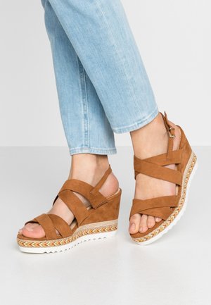 Feet wearing brown suede wedge sandals with crisscross straps and buckle, paired with light blue denim jeans against a plain background.