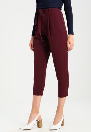 Vrouw die cropped bordeauxrood paperbag-taillepants met een gestrikte riem draagt en witte slingback schoenen met blokhak tegen een witte achtergrond.