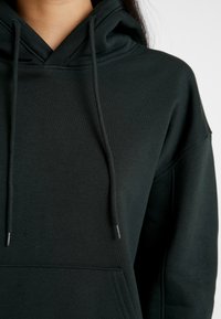 Tmavě zelená mikina, vyrobená z měkké látky, má klokaní kapsu, šňůrky na stažení a klasický oversized střih s žebrovanými manžetami.