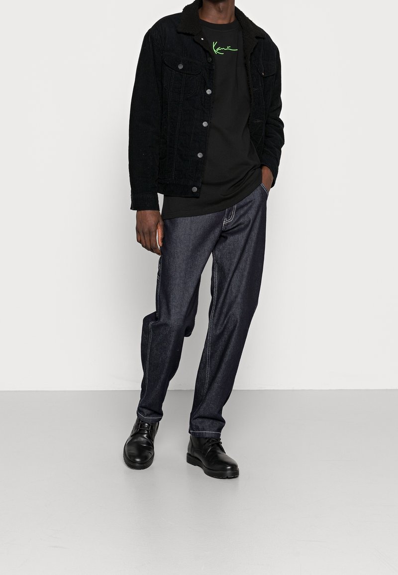 Černá kordurová bunda s límcem z sherpy, nošená přes černé tričko s zelenou grafikou, doplněná tmavými džínovými kalhotami a černými botami.