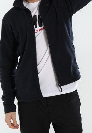 Person iført en delvist lynlåst sort jakke over en hvid t-shirt med grafik og sorte bukser, stående foran en ensfarvet baggrund.