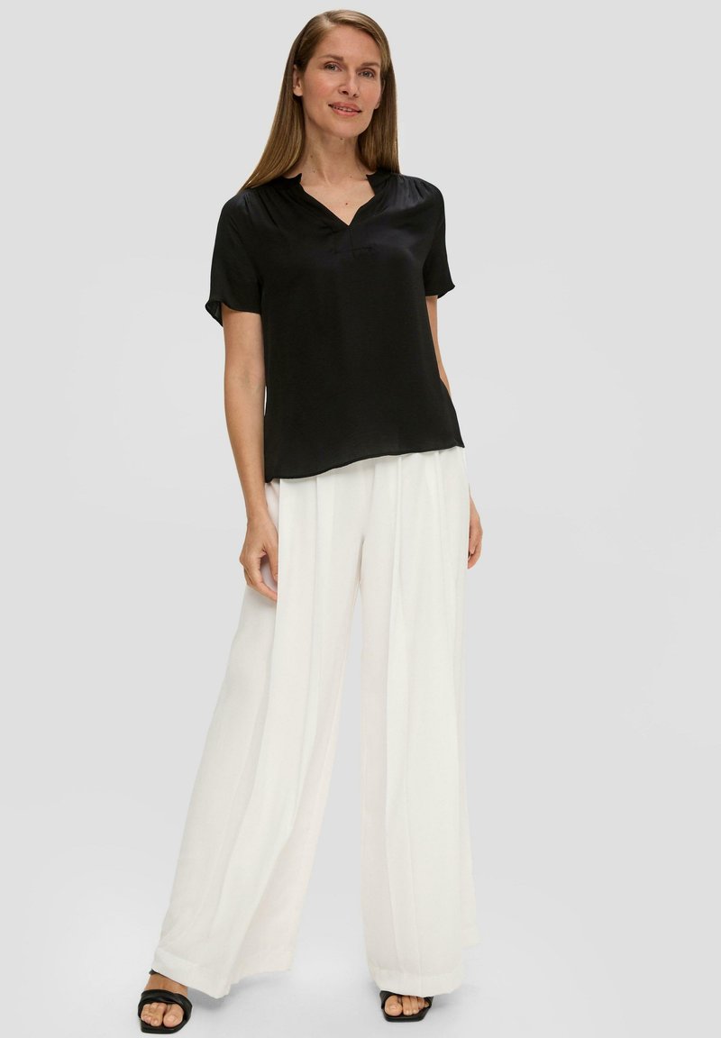 Zwarte blouse met korte mouwen en een V-hals, gecombineerd met witte wijde broekspijpen. De broek heeft een geplooid ontwerp en een luchtige textuur.