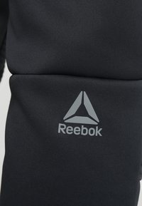 Svart idrottstyg med en mjuk textur som har en grå Reebok-logotyp och en triangulär ikon. Designen är stilren och minimalistisk.