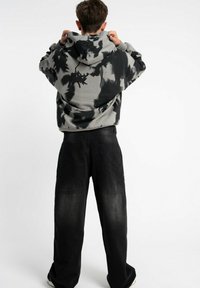 Osoba widziana z tyłu, nosząca czarną i szaro-tie-dye bluzę z kapturem oraz luźne czarne spodnie, poprawiająca kaptur.