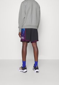 Sudadera gris con puños acanalados, pantalones cortos negros con ribetes morados y un panel lateral colorido, calcetines azules y zapatillas blancas/negras con acentos dorados.