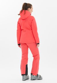 Helle Koralle-Ski-Ausrüstung mit Kapuze, bestehend aus einer taillierten Jacke und lockeren Hosen. Beinhaltet schwarze Akzente und graue Skischuhe. Glatte Textur.