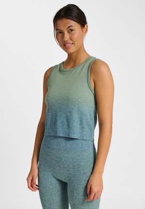 Ung kvinde stående iført ærmeløs grøn og blå ombre top med matchende højtaljede leggings mod ensfarvet hvid baggrund.