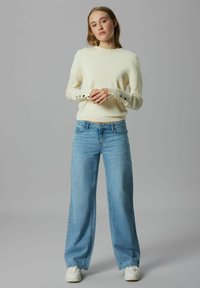 Kremowy sweter z guzikami na mankietach, zestawiony z jasnoniebieskimi szerokimi jeansami i białymi trampkami. Prosty i casualowy design.