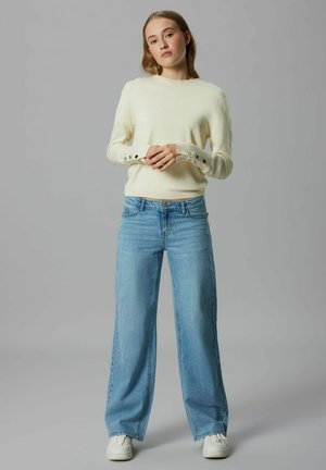 Džemper krem boje s gumbima na manšetama, uparen s svijetloplavim trapericama širokih nogavica i bijelim tenisicama. Jednostavan i ležeran dizajn.