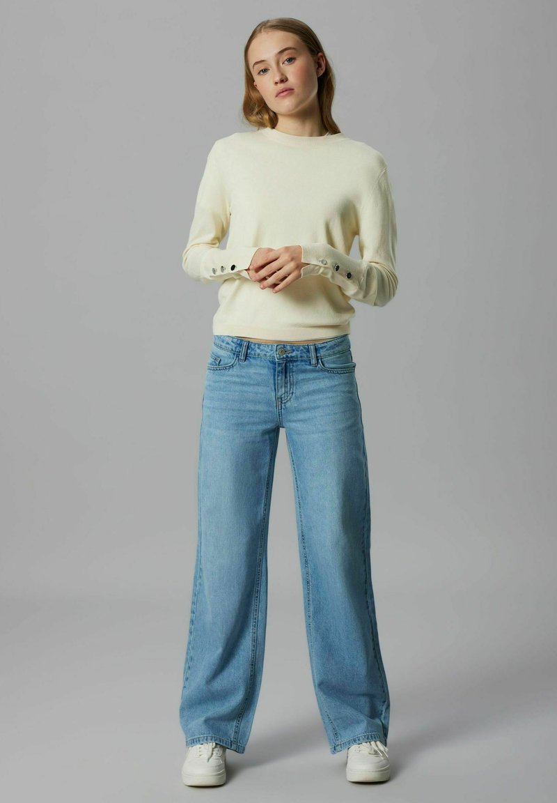 Kremowy sweter z guzikami na mankietach, zestawiony z jasnoniebieskimi szerokimi jeansami i białymi trampkami. Prosty i casualowy design.