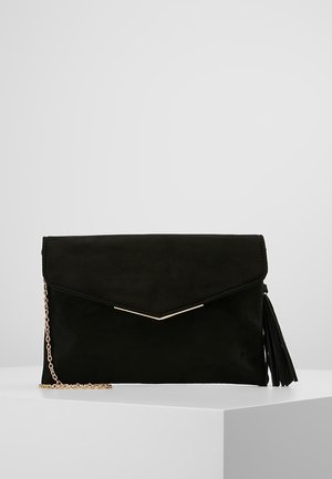 Schwarze Clutch mit goldener Kettenriemen und Quasten-Detail, auf einer weißen minimalistischen Oberfläche vor hellem Hintergrund liegend.
