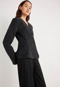 Čierne priliehavé sako z mäkkej tkaniny s výstrihom do V a piatimi gombíkmi. Dlhé rukávy, priliehavý strih a mierne rozšírený spodný lem.