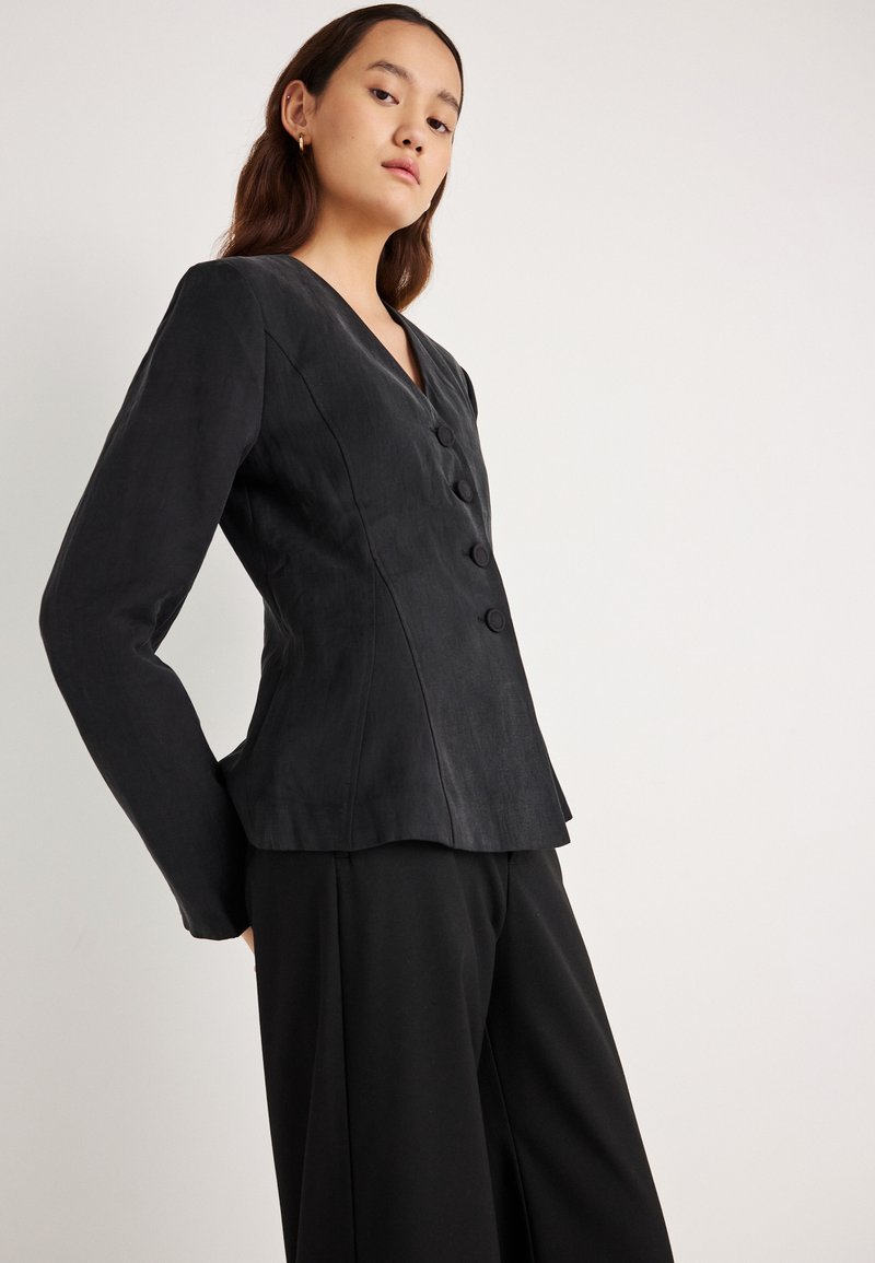 Čierne priliehavé sako z mäkkej tkaniny s výstrihom do V a piatimi gombíkmi. Dlhé rukávy, priliehavý strih a mierne rozšírený spodný lem.
