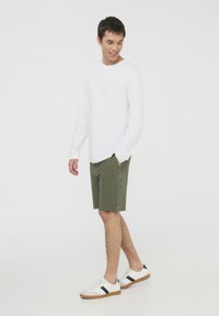 Camicia bianca a maniche lunghe, pantaloni corti verde oliva con stampa del logo laterale, scarpe da ginnastica bianche con strisce nere. Outfit semplice e casual con una vestibilità rilassata.