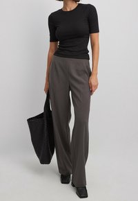 Zwarte geribbelde top met korte mouwen gecombineerd met hooggesneden grijze geklede broek. Model houdt een geweven zwarte tote bag vast. Vierkante zwarte schoenen.