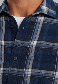 Blauwe en witte geruite flanellen blouse met knopen, voorzien van een borstzak en een label met de tekst "PME Legend." Close-up van de textuur van de stof.