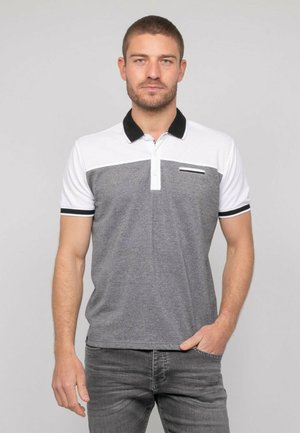 Homme debout, portant un polo à manches courtes gris et blanc avec un col noir et un jean gris, la main partiellement dans la poche.