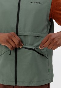 Gilet leggero verde con tasca con zip. Presenta una zip frontale e un cordino, realizzato in tessuto liscio e opaco. Logo Vaude visibile.