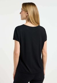 Zwart短袖top met een ontspannen pasvorm. De stof heeft een zachte textuur. Heeft een ronde halslijn aan de achterkant. Draagt met zwarte broek.