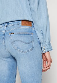 Jeans de mezclilla azul claro con una parche de cuero de Lee en la cintura. Presenta bolsillos traseros con costuras en curva y una textura suave.