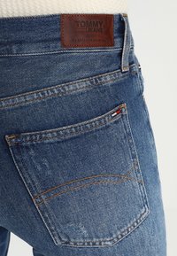 Jeans de mezclilla con un lavado azul, que cuentan con una etiqueta de cuero marrón con la inscripción "TOMMY JEANS", detalles de costura y un pequeño ancla en el bolsillo lateral.