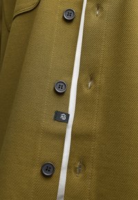 Olivengrøn frakke med struktureret stof, sorte knapper, hvid accentstribe og et lille sort label med et logo nær sidesømmen.