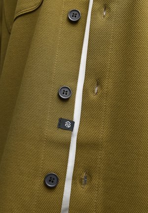 Olivgrüner Mantel mit strukturierter Stoffoberfläche, schwarzen Knöpfen, einem weißen Akzentstreifen und einem kleinen schwarzen Etikett mit Logo in der Nähe der Seitennaht.