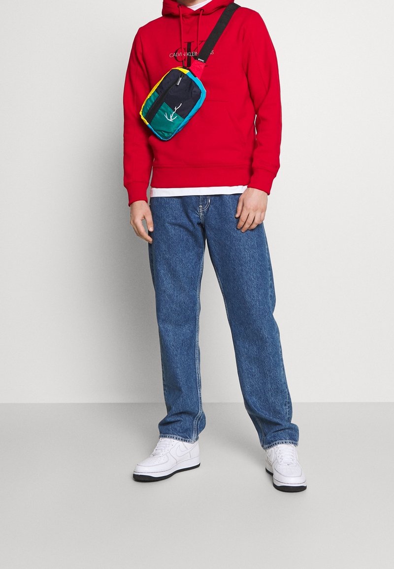 Personne portant un sweat à capuche rouge Calvin Klein, un jean bleu, des baskets blanches et un sac bandoulière multicolore avec une sangle noire.