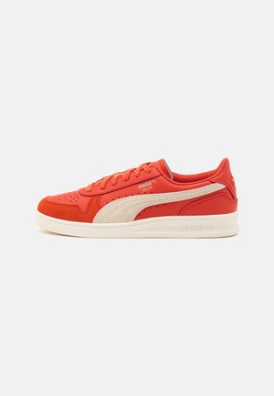 Zapatilla deportiva en rojo y beige con costados perforados, cuello acolchado, cordones planos y suela de goma texturizada. Logo de Puma visible.
