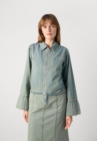 Kvinne som har på seg en lyseblå denimjakke med vide ermer og matchende denimskjørt, står mot en ensfarget lys bakgrunn.