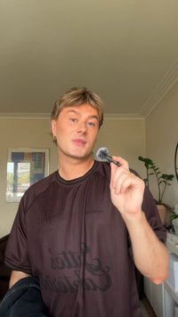 Schwarzer Make-up-Pinsel mit einer flauschigen, grauen Spitze, vor einem Hintergrund aus Zimmerpflanzen und gerahmter Kunst; trägt ein dunkelbraunes Hemd.