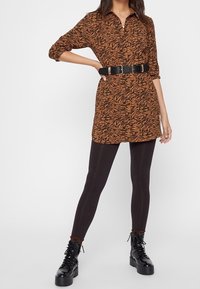 Vestito camicia con stampa animale marrone e nera, colletto, apertura a bottoni frontale, fasciante in vita con una cintura nera, abbinato a leggings neri e stivali.