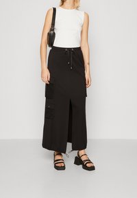 Mujer con top blanco sin mangas, falda larga negra con abertura frontal y bolsillos, sandalias de tacón negras y bolso negro de hombro.