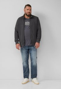 Man met een baard in een donkergrijze zip-up jas, een grijs T-shirt met print, blauwe jeans en witte sneakers, staand tegen een witte achtergrond.