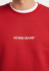 Rød hettegenser med ribbestrikket mannsstrikket hals. Har hvit brodert "GUESS JEANS"-logo på brystet. Stoffblanding av bomull, glatt tekstur.