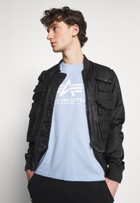 Černá bomber bunda s několika předními kapsami, hladkou texturou a žebrovanými manžetami. Nošena přes světle modré tričko s grafickým logem.