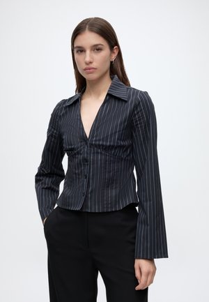 Femme portant une chemise noire à fines rayures boutonnée avec manches longues et un pantalon noir, debout avec une main dans la poche sur fond blanc.