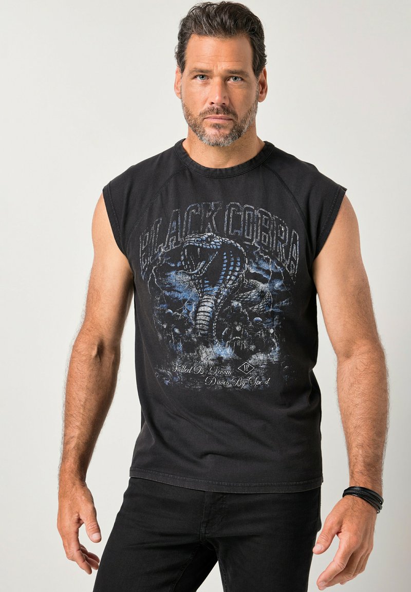 Man met kort donker haar en baard, gekleed in een mouwloos zwart shirt met een blauwe "Black Cobra"-afbeelding en een zwarte broek, staand tegen een effen achtergrond.