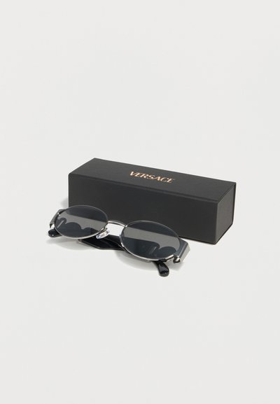 Οβάλ μαύρα γυαλιά ηλίου με ασημένιο σκελετό που ακουμπούν δίπλα σε μία μαύρη ορθογώνια θήκη με το λογότυπο της Versace σε λευκό φόντο.