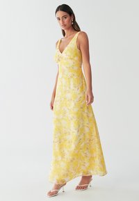 Žlté maxi šaty s kvetinovým vzorom, hlbokým výstrihom do V, nastaviteľnými ramienkami, ľahkou látkou a prúdiacim strihom. Kombinované s bielymi sandálmi.