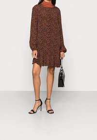 Kleid mit braunem Leopardenmuster, langen Ärmeln, fließendem Schnitt und geraffter Textur. Kombiniert mit schwarzen Riemchen-Heels und einer schwarzen Handtasche.