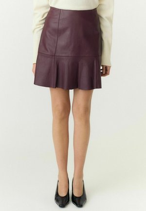 Femme portant une mini-jupe en cuir bordeaux avec des plis et des talons noirs pointus, debout devant un fond uni.