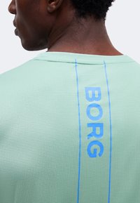 Κοντινή εικόνα ενός ατόμου που φοράει ένα ανοιχτοπράσινο υφασμάτινο πουκάμισο με κάθετο μπλε κείμενο «BORG» στο πίσω μέρος κοντά στο λαιμό.