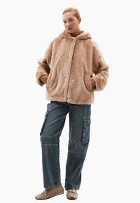 Beige bunda s kapucí z umělé kožešiny, velkými předními kapsami, stříbrnými knoflíky a volným střihem, spárovaná s modrými cargo džíny.