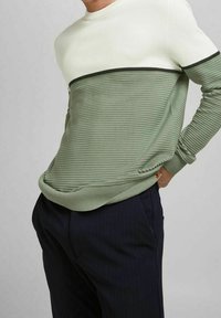 Man in een ribgebreide trui met een witte top en een olijfgroene onderkant, gedragen met donkerblauwe broek, hand in zak, tegen een effen achtergrond.
